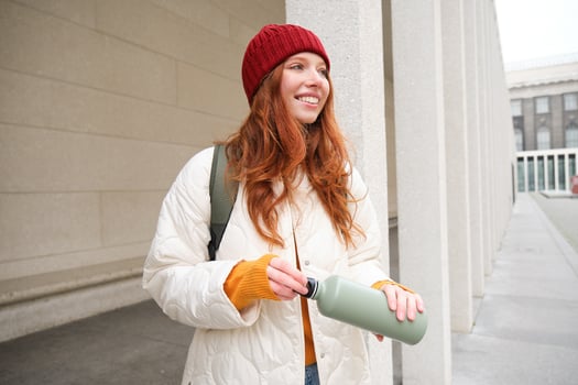 Young happy redhead woman in red hat, drinking from thermos, warming up with hot drink in her flask while walking around city, tourist relaxes with warm refreshemtn photo