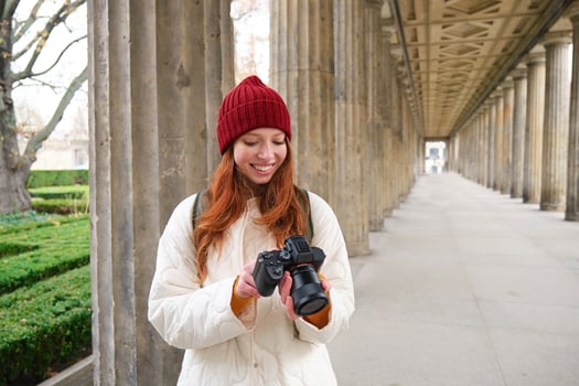Young redhead female photographer, makes lifestyle shooting in city centre, takes photos and smiles, looks for perfect shot, makes picture