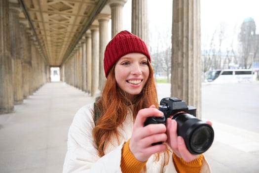 Young redhead female photographer, makes lifestyle shooting in city centre, takes photos and smiles, looks for perfect shot, makes picture