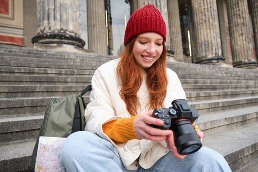 Young student, photographer sits on street stairs and checks her shots on professional camera, taking photos outdoors