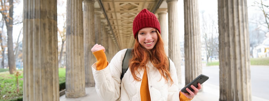 Smiling redhead girl looks excited, tourist holding mobile phone and does fist pump, thrilled about her journey, using smartphone on street photo