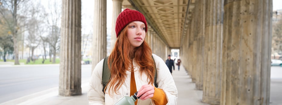 Young redhead girl holds thermos in hands, pours herself a hot drink while walking in city. Tourist takes break, opens flask for refreshment beverage photo