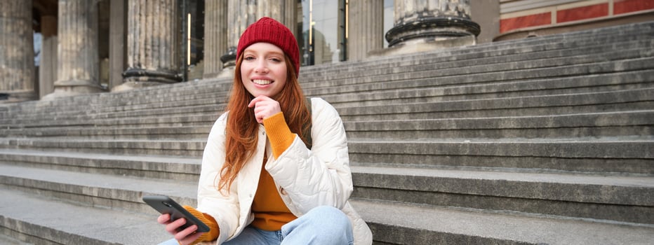 Stylish young redhead woman, talking on mobile phone app, using social media application, looking for something online on smartphone, sits on stairs outdoors photo