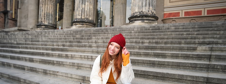 Stylish young redhead woman, talking on mobile phone app, using social media application, looking for something online on smartphone, sits on stairs outdoors photo