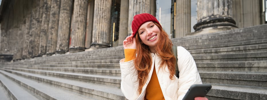 Portrait of young redhead woman in red hat, sitting on stairs, tourist looks at her mobile phone, rests on staircase of museum, connects public wifi photo