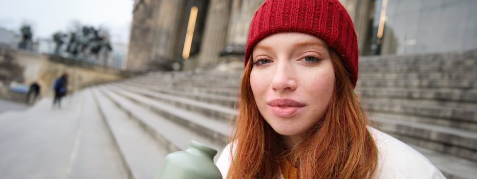 Young redhead female tourist rests during her trip, opens thermos and drinks hot tea, having a break after sightseeing photo