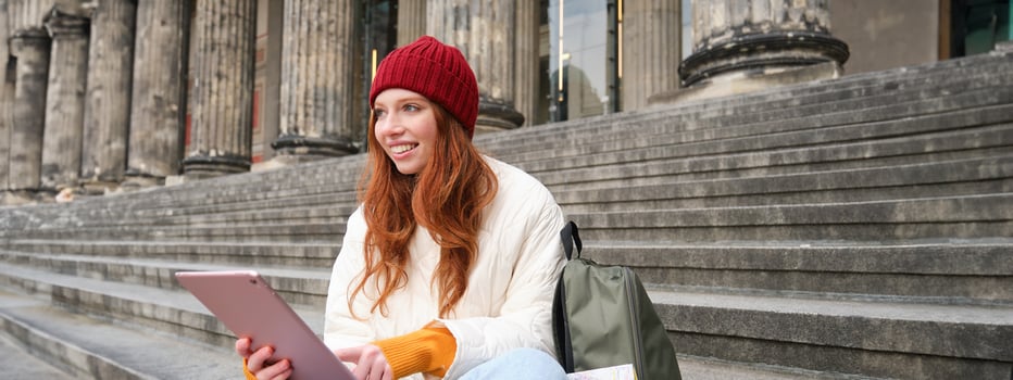 Beautiful young modern girl with red hair, holds digital tablet, sits on stairs near museum and connects public internet, sends message on gadget app photo