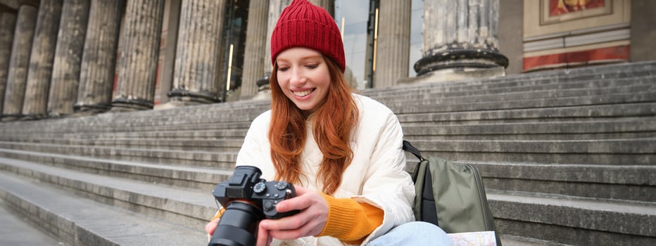 Young student, photographer sits on street stairs and checks her shots on professional camera, taking photos outdoors