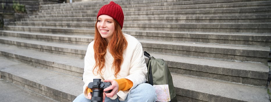 Young student, photographer sits on street stairs and checks her shots on professional camera, taking photos outdoors