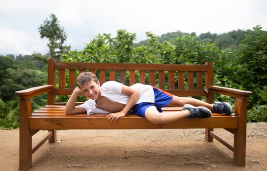A happy child in the summer heat lies on a wooden bench against the backdrop of tropical plants. The boy is on vacation in the summer. photo