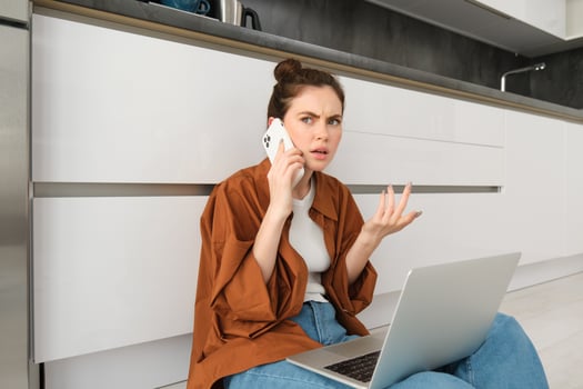 Woman with frustrated and confused face talking on mobile phone, shrugging and frowning, hear bad news over the telephone, sits on floor with laptop, calls customer support photo