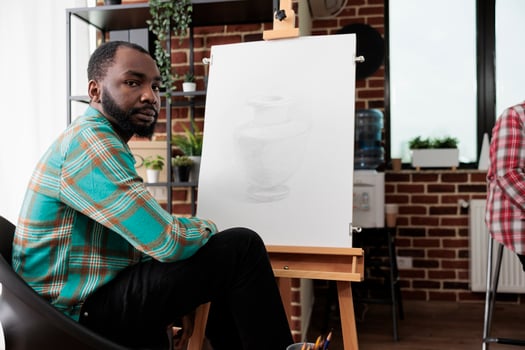 African American guy sitting at easel in classroom and looking at camera, studying fine art. People participating in drawing masterclass, learning various techniques centred around pencil sketching. photo