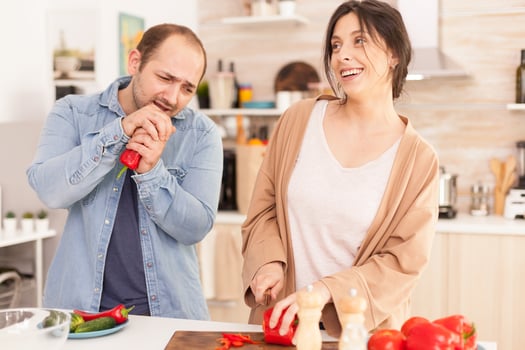 Man singing on red pepper in kitchen making wife smile. Girlfriend preparing salad. Funny happy in love couple at home spending time together, cooking healthy and smiling photo
