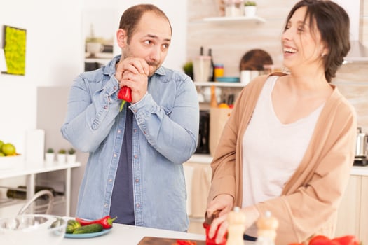 Husband looking at wife while she is smiling because of him in kitchen. Cutting red pepper on wooden chopper. Funny happy in love couple at home spending time together, cooking healthy and smiling photo