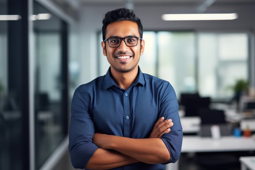 AI generated a young Indian male designer, engineer, architect who is wearing glasses and a blue shirt smiling standing in the office and looking at the camera photo