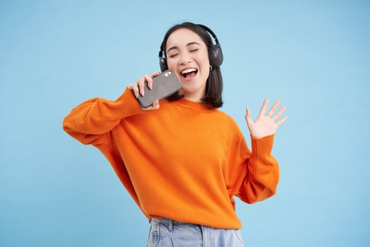 Beautiful korean girl singing in smartphone mic, listens music in headphones, sings karaoke on mobile app, stands over blue background photo