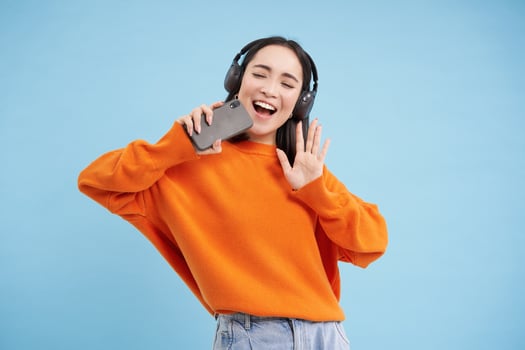 Beautiful korean girl singing in smartphone mic, listens music in headphones, sings karaoke on mobile app, stands over blue background photo