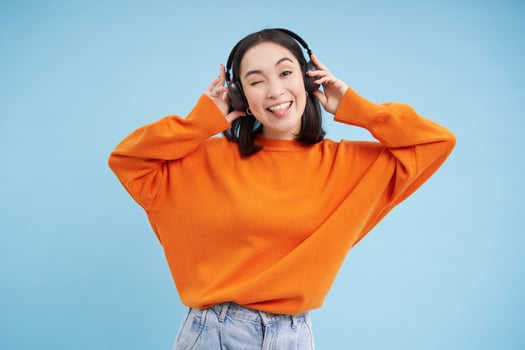 Happy Chinese woman in headphones, listens music, enjoys favourite song in her playlist, stands over blue background photo