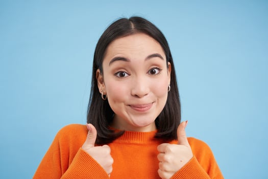 Close up portrait of asian woman shows thumbs up, smiles and looks supportive, approves, recommends smth, gives positive reply, blue background photo