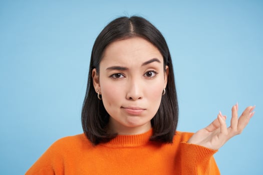 So what. Portrait of korean woman shrugs and raises arm up with skeptical, confused face expression, stands over blue background photo