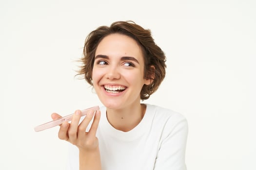 Laughing, happy woman talking on speakerphone, holding phone near mouth, recording voice message on smartphone, standing over white background photo