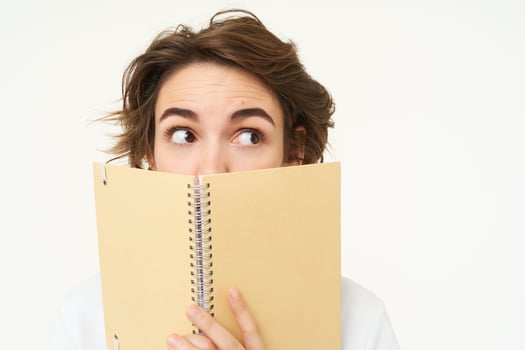 Woman of cute brunette woman hiding her face behind planner, peeking aside, looking right at banner, standing over white background photo