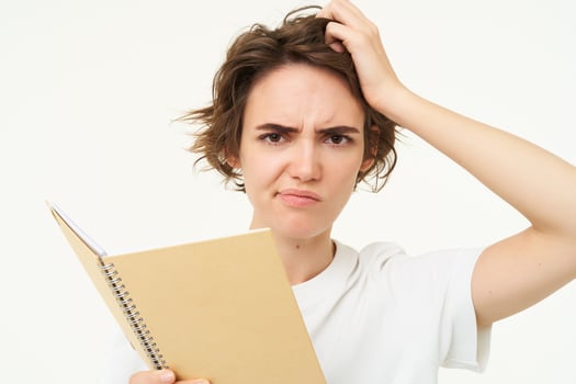 Portrait of confused girl scratching her head, holding planner, student doing homework with puzzled face, thinking with frowning expression, standing over white background photo