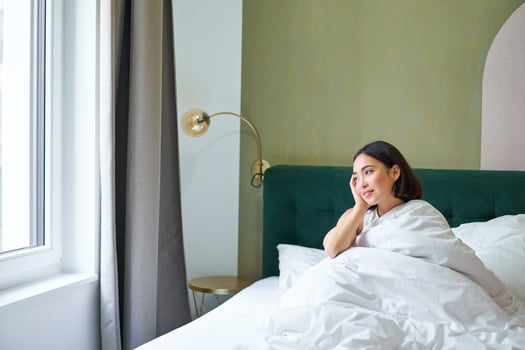 Happy korean woman wakes up in her hotel room, lying in bed under cozy warm blankets, white sheets, enjoying weekend morning photo