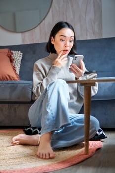 Vertical shot of young asian girl looks surprised at her mobile phone, reads amazing news, notification on smartphone app, sits at home on floor photo