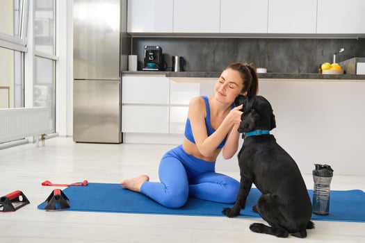 Portrait of beautiful brunette fitness girl, plays with her dog during workout training session, sits at home on rubber yoga mat, does sport exercises in living room photo