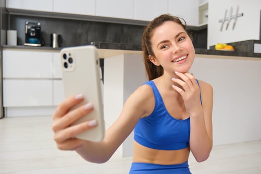 Portrait of young fitness girl, blogger records her workout from home, poses for selfie on smartphone, does yoga on camera, sits in blue sportsbra and leggings photo