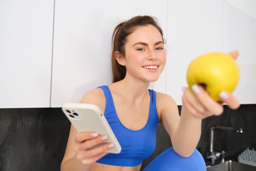 Image of beautiful, sportswoman sitting in kitchen with smartphone, offering you an apple, eating fruits, wearing sportsbra and leggings photo