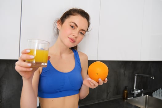 Close up portrait of sportswoman, fitness girl holding glass of fresh juice and an orange in hands, smiling at camera, standing in kitchen photo