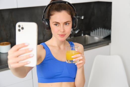 Portrait of young lifestyle, fitness blogger, woman with orange juice, takes selfie on smartphone camera, posing in activewear and wireless headphones photo