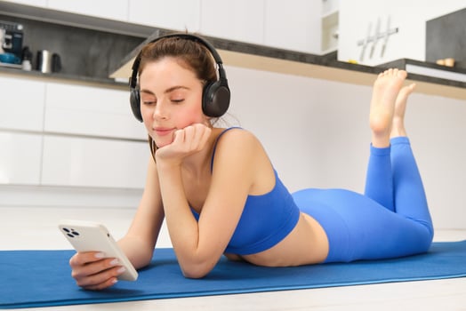 Portrait of brunette fitness girl, laying on yoga mat at home, watching workout tutorials, videos on smartphone, wearing sportswear and wireless headphones photo
