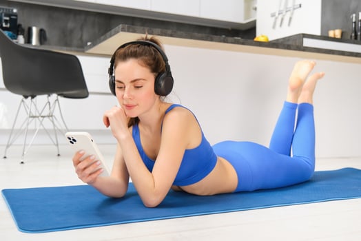 Image of young woman watches sport gym tutorials on smartphone, laying on yoga mat, doing fitness workout at home, wearing blue leggings and sportsbra photo