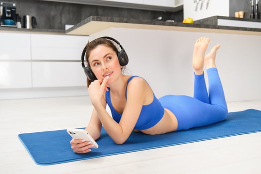 Image of smiling young woman in fitness clothing, listening music in wireless headphones, holding smartphone, using sports app while doing workout at home photo