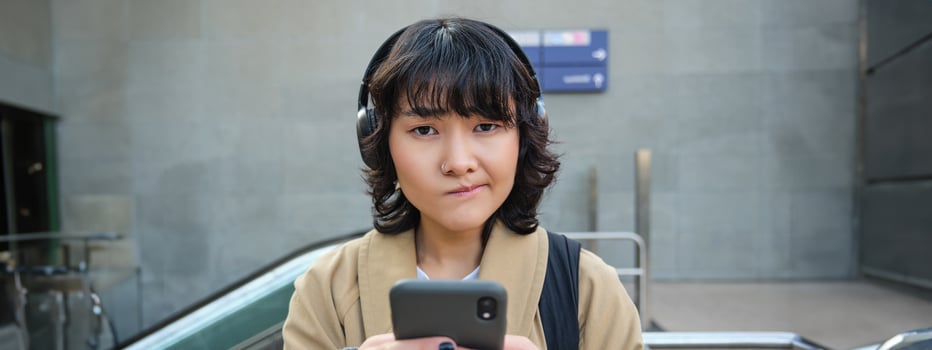 Close up portrait of puzzled, complicated korean girl in headphones, frowns and looks unsure, stands with smartphone and looks at camera while shrugs on street photo