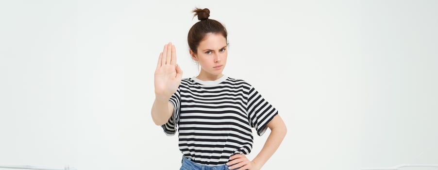 Image of beautiful woman with serious face, showing stop sign, extends one palm and frowns, stands over white background photo