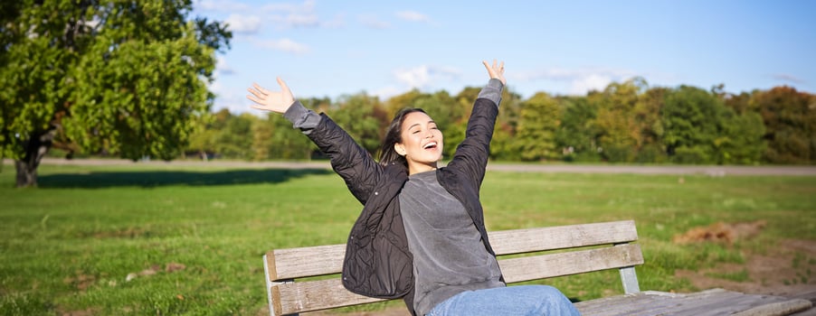 Happy asian woman stretching her hands, sitting on bench with excited face, smiling pleased, feeling freedom, enjoying day in park photo