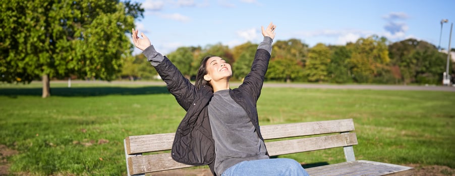 Happy asian woman stretching her hands, sitting on bench with excited face, smiling pleased, feeling freedom, enjoying day in park photo