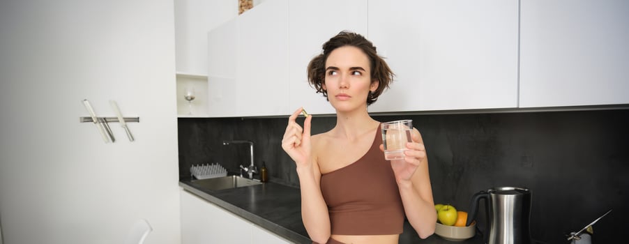 Portrait of sportswoman drinking water, taking vitamins, dietary supplements for healthy skin, having omega-3 pill, standing in her kitchen in workout clothing photo