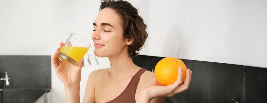 Healthy lifestyle and sport. Beautiful smiling woman, drinking fresh orange juice and holding fruit in her hand, enjoying her vitamin drink after workout at home, standing in kitchen photo