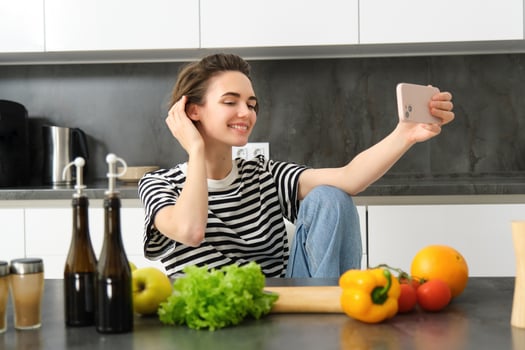 Cute young woman, modern lifestyle blogger, taking selfie while cooking salad in the kitchen, using smartphone to make photos for social media app