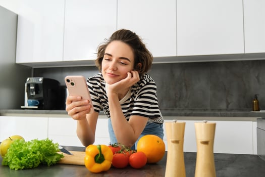 Portrait of beautiful young woman with smartphone, cooking in the kitchen, chopping vegetables, reading recipe online photo