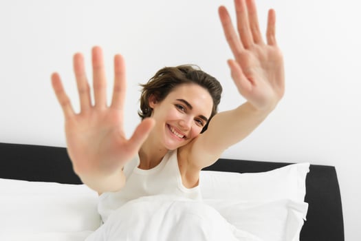 Beautiful young woman stretching her hands towards camera, waking up in morning, resting in her bed, smiling and looking happy photo