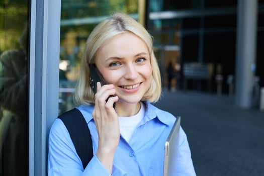 Close up portrait of modern young woman, student standing near building in city centre, talking on mobile phone, having chat on smartphone and smiling photo