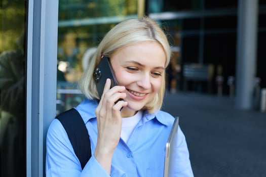 Happy female model, blond woman talking on mobile phone, chatting with someone, standing near office building with laptop and smartphone, smiling photo