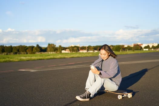 Portrait of young korean girl sitting on her skateboard on road, looking at smartphone, chatting on mobile app photo