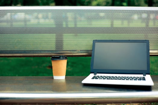 Mockup image of laptop with blank black screen and coffee cup on metal bench in nature outdoor park photo